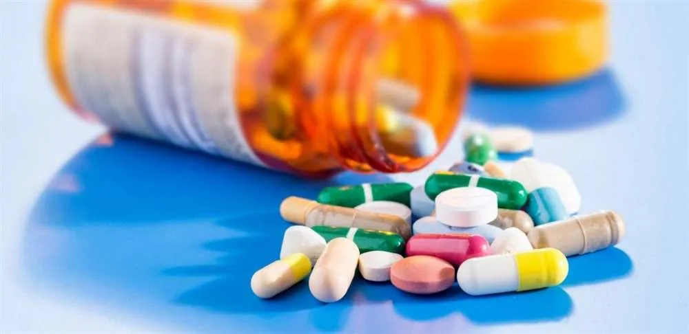 An open pill bottle spilling a variety of over-the-counter tablets and capsules onto a blue surface